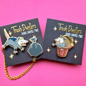 Two sets of hard enamel pins with gold plating. The left set depicts a possum pin with wide stance and an open mouth, and is connected with a loose gold chain to a pin depicting a trashbag. The right pin is a yelling possum in a golden trash can.