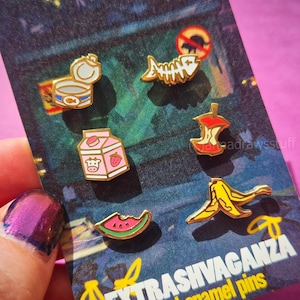 May include: A set of six gold-toned enamel pins displayed on a backing card labeled EXTRASHVAGANZA. The pins feature designs of an open tin can, a fish skeleton, a strawberry milk carton, an apple core, a watermelon slice, and a banana peel.