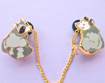 May include: A gold-toned metal enamel pin set featuring a raccoon and an opossum connected by a delicate gold chain. The raccoon has grey and white fur with a black mask, while the opossum has white fur, a pink nose, and a pink tail.