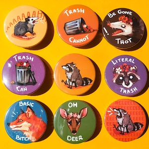 May include: Nine round pins with colorful illustrations and text. The pins feature a raccoon, a deer, a fox, a trash can, and a possum. The text on the pins includes "Trash", "Literal Trash", "Be Gone Thot", "Oh Deer", "Basic Bitch", "AAAAAH", "Trash Cannot", and "Reeeeeeee".