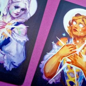 Puede incluir: Dos ilustraciones de mujeres con luz blanca brillante que emana de sus manos. La mujer de la izquierda tiene el pelo corto y blanco y lleva un vestido morado y blanco. La mujer de la derecha tiene el pelo largo y rubio y lleva un vestido blanco y amarillo. Ambas mujeres tienen halos blancos alrededor de la cabeza.