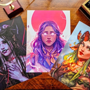 Op de afbeelding: Drie fantasy kunstprints met portretten van mystieke figuren. Eén print toont een vrouw met lang zwart en wit haar, een andere toont een vrouw met paars haar en een roze zon, en de derde toont een vrouw met hoorns en groen haar.