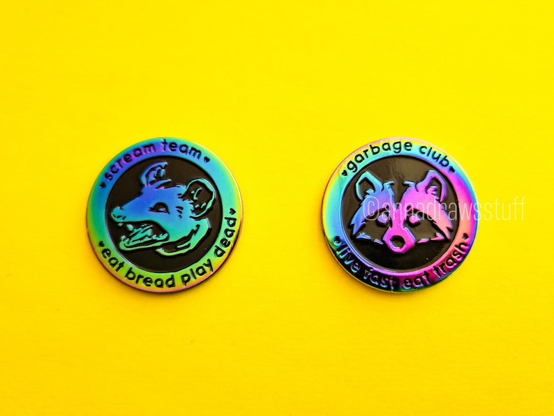 May include: Two iridescent enamel pins with black backgrounds. The first pin features a cartoon opossum with the text "scream team. eat bread play dead". The second pin features a cartoon raccoon with the text "garbage club. live fast eat trash".