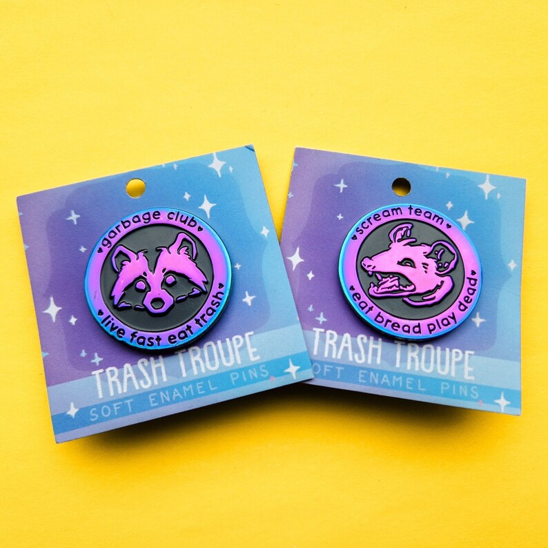 Op de afbeelding: Twee emaillepins met een zwarte en iriserende afwerking. De eerste pin heeft een wasbeer met de tekst "garbage club. live fast eat trash". De tweede pin heeft een opossum met de tekst "scream team. eat bread play dead". Beide pins hebben de tekst "TRASH TROUPE SOFT ENAMEL PINS".