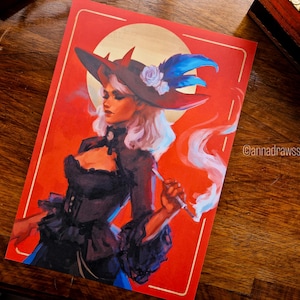 Puede incluir: Impresión de una mujer con sombrero de bruja, fumando, sobre un fondo rojo con un círculo dorado. La mujer lleva un vestido negro y un sombrero con cuernos, plumas y una flor. El nombre del artista, @annadrawsstuff, es visible.
