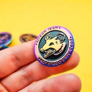 Op de afbeelding: Een regenboogkleurige, iriserende emaille pin met een zwarte en gouden illustratie van een opossumkop met de tekst "scream team eat bread play dead".