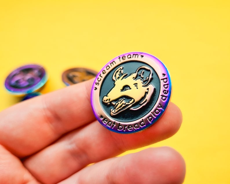 Op de afbeelding: Een regenboogkleurige, iriserende emaille pin met een zwarte en gouden illustratie van een opossumkop met de tekst "scream team eat bread play dead".