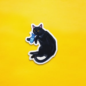 May include: A black cat with yellow eyes is holding a blue fish toy in its mouth. The cat is sitting on a yellow background.