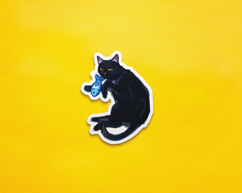 May include: A black cat with yellow eyes is holding a blue fish toy in its mouth. The cat is sitting on a yellow background.