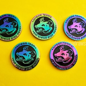 Op de afbeelding: Vijf iriserende emaillepins met een zwarte achtergrond en een afbeelding van een cartoondier met scherpe tanden. De tekst op de pins luidt "scream team. eat bread play dead".