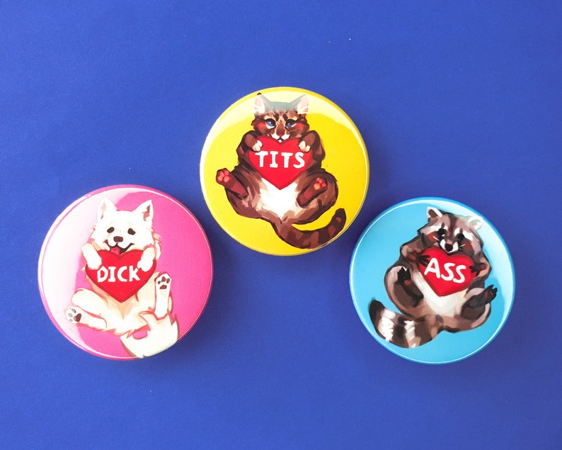 May include: Three round button pins with cartoon animals holding hearts with different words on them. The first pin has a white dog holding a heart that says "DICK". The second pin has a brown and white cat holding a heart that says "TITS". The third pin has a raccoon holding a heart that says "ASS".