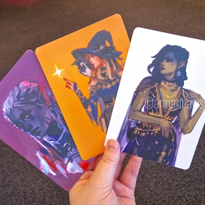 Puede incluir: Tres cartas ilustradas con personajes de fantas&iacute;a. La primera carta es morada con un retrato de una persona con piel azul y cabello oscuro. La segunda carta es naranja con una persona que lleva un sombrero de bruja y sostiene una varita. La tercera carta es blanca con una persona con cabello largo y oscuro que lleva un atuendo morado y dorado. El texto en la tercera carta dice "@cannadrasstuff".
