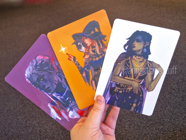 Puede incluir: Tres cartas ilustradas con personajes de fantas&iacute;a. La primera carta es morada con un retrato de una persona con piel azul y cabello oscuro. La segunda carta es naranja con una persona que lleva un sombrero de bruja y sostiene una varita. La tercera carta es blanca con una persona con cabello largo y oscuro que lleva un atuendo morado y dorado. El texto en la tercera carta dice "@cannadrasstuff".