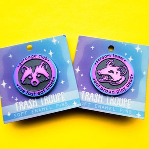 May include: Two enamel pins with a black and purple iridescent finish. The first pin features a raccoon with the text "garbage club, live fast eat trash". The second pin features a possum with the text "scream team, eat bread play dead". Both pins have the text "TRASH TROUPE SOFT ENAMEL PINS" on the card.