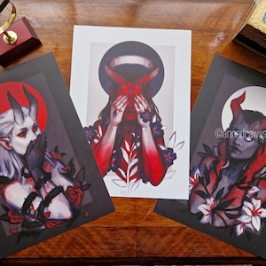 May include: Three art prints featuring demonic figures with horns, set against dark backgrounds. The central print shows a figure covering their eyes, while the others depict figures with horns and floral accents. The artwork has a gothic theme.