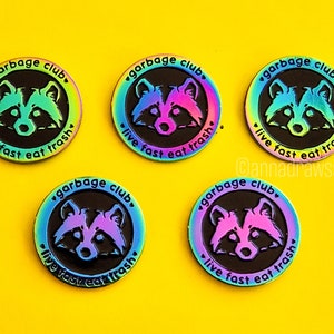 Op de afbeelding: Vijf regenboogkleurige emaillepins met een wasbeer en de tekst "garbage club, live fast eat trash".