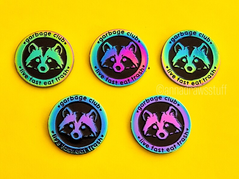 Op de afbeelding: Vijf regenboogkleurige emaillepins met een wasbeer en de tekst "garbage club, live fast eat trash".