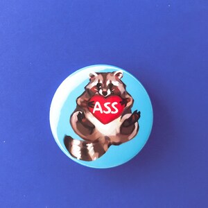 May include: A blue button with a cartoon raccoon holding a red heart with the word "ASS" on it.