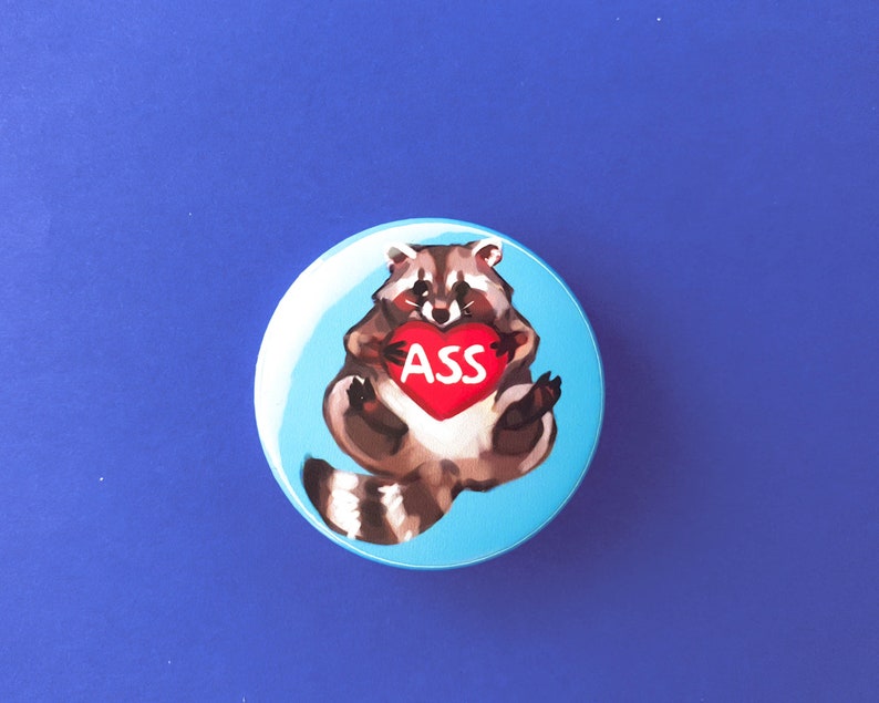 May include: A blue button with a cartoon raccoon holding a red heart with the word "ASS" on it.