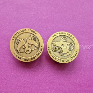 Puede incluir: Dos pines de color dorado con ilustraciones de animales. El primer pin presenta un mapache con el texto "garbage club. live fast eat trash". El segundo pin presenta una rata con el texto "scream team. eat bread play dead".