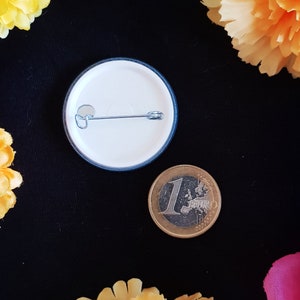 May include: The back of a white button with a blue border. The button has a safety pin attached to it. A euro coin is next to the button.