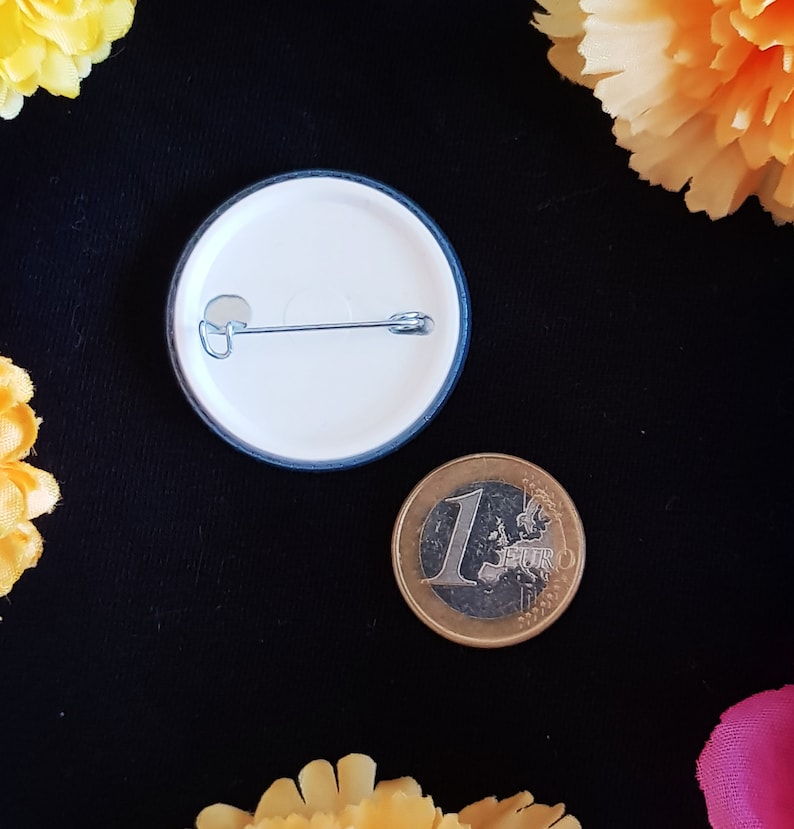 May include: The back of a white button with a blue border. The button has a safety pin attached to it. A euro coin is next to the button.