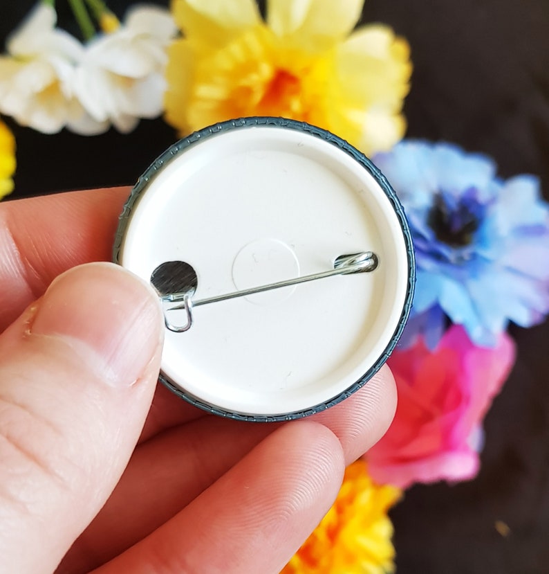 May include: The back of a round, black and white button with a silver safety pin attached. The button has a white center and a black outer ring.