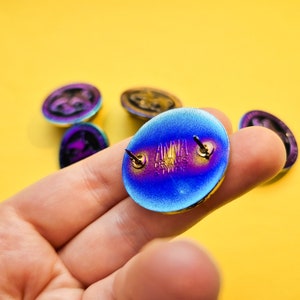 Op de afbeelding: Een close-up van de achterkant van een blauwe, paarse en gouden iriserende emaille pin. De pin heeft een metalen pen met een vlinder sluiting. De pin heeft het woord "ANIMAL" erop geschreven.