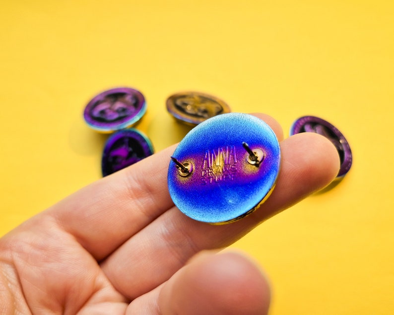 Op de afbeelding: Een close-up van de achterkant van een blauwe, paarse en gouden iriserende emaille pin. De pin heeft een metalen pen met een vlinder sluiting. De pin heeft het woord "ANIMAL" erop geschreven.