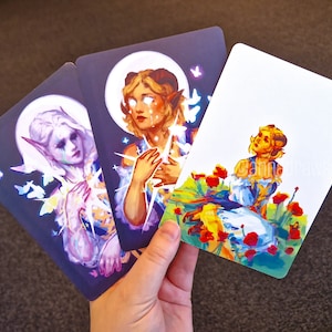 Puede incluir: Tres cartas ilustradas con personajes de fantas&iacute;a sobre fondos coloridos. La primera carta presenta un personaje con cabello morado con una corona de flores y mariposas. La segunda carta presenta un personaje con cabello rubio y una corona de estrellas. La tercera carta presenta un personaje con cabello rubio y una corona de flores en un campo de flores rojas.