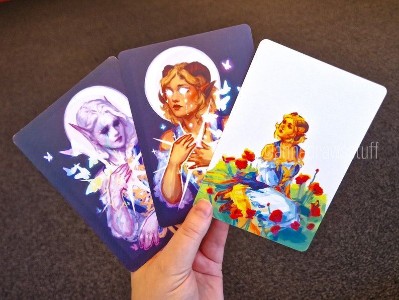 Puede incluir: Tres cartas ilustradas con personajes de fantas&iacute;a sobre fondos coloridos. La primera carta presenta un personaje con cabello morado con una corona de flores y mariposas. La segunda carta presenta un personaje con cabello rubio y una corona de estrellas. La tercera carta presenta un personaje con cabello rubio y una corona de flores en un campo de flores rojas.