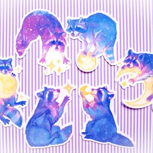 Puede incluir: Un conjunto de pegatinas de mapaches caprichosos con un tema de galaxia. Los mapaches están representados en tonos de azul, morado y rosa, interactuando con lunas y estrellas. Las pegatinas están sobre un fondo a rayas morado y blanco.
