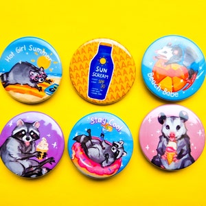 Könnte beinhalten: Sechs bunte runde Pins mit verschiedenen Designs, die Waschbären und Opossums zeigen. Die Pins haben einen weißen Hintergrund mit bunten Illustrationen und Text. Der Text auf den Pins lautet "Hot Girl Summer", "Beach Babe", "Stay Cool" und "Sun Scream SPF 30".