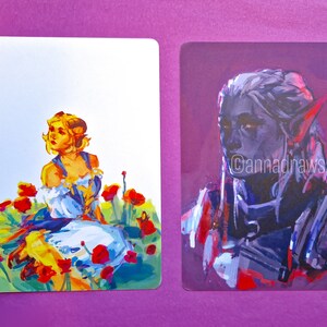 Puede incluir: Dos ilustraciones pintadas sobre cartulina blanca. La ilustraci&oacute;n de la izquierda muestra a una mujer con cabello rubio y un vestido blanco sentada en un campo de amapolas rojas. La ilustraci&oacute;n de la derecha muestra un retrato de una persona con cabello gris y piel azul. El texto "@cannadrasstuff" est&aacute; en la esquina inferior derecha de la ilustraci&oacute;n.