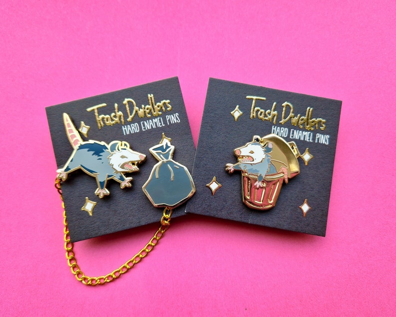 Two sets of hard enamel pins with gold plating. The left set depicts a possum pin with wide stance and an open mouth, and is connected with a loose gold chain to a pin depicting a trashbag. The right pin is a yelling possum in a golden trash can.