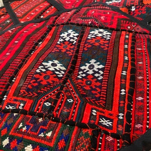 Op de afbeelding: Een rood, zwart en wit gepatroneerd textiel met ingewikkelde geometrische ontwerpen. Het textiel is gemaakt van geweven stof en heeft een ruwe textuur. Het textiel is een traditioneel ontwerp en is waarschijnlijk afkomstig uit een land in het Midden-Oosten of Azië.