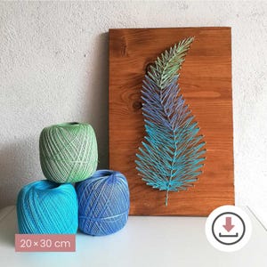 Könnte beinhalten: Ein Wandkunstwerk aus Holz mit einem Federdesign aus Faden in Blau- und Grüntönen. Drei Garnrollen in Grün, Blau und Türkis sind vor der Kunst gestapelt. Das Kunstwerk misst 20 x 30 cm.