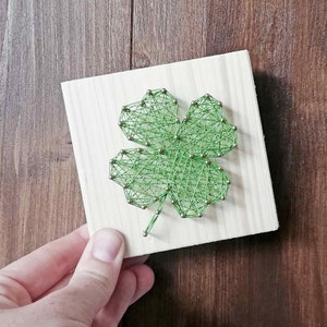 Könnte beinhalten: Grüne String Art eines vierblättrigen Kleeblatts auf einem Holzgrund.