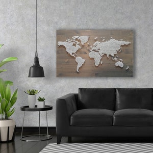 Könnte beinhalten: Eine Weltkarte aus weißen Stecknadeln auf braunem Holzgrund. Die Karte hängt an einer Wand über einem schwarzen Ledersofa und einem schwarzen Beistelltisch mit zwei Topfpflanzen.