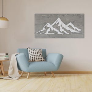 Könnte beinhalten: Graue Wandkunst aus Holz mit einem weißen Bergketten-Design. Das rechteckige Kunstwerk ist auf einer hellgrauen Holzoberfläche montiert. Das Bild wird über einem blauen Sessel in einem Wohnzimmer gezeigt.