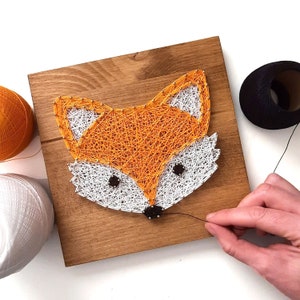 Könnte beinhalten: Ein Holzrahmen mit einem String-Art-Fuchsdesign. Der Fuchs ist orange und weiß mit schwarzen Augen und einer schwarzen Nase. Der Rahmen wird mit schwarzem Garn bearbeitet.
