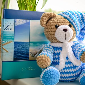 May include: A crocheted teddy bear wearing a blue and white striped sleepwear outfit and a matching blue and white hat. The bear is sitting on a windowsill next to a photo album with a sea theme.
