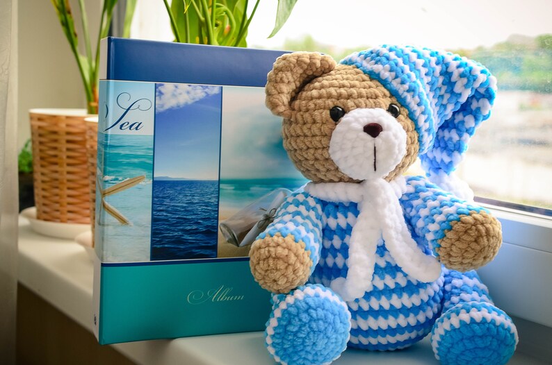 May include: A crocheted teddy bear wearing a blue and white striped sleepwear outfit and a matching blue and white hat. The bear is sitting on a windowsill next to a photo album with a sea theme.
