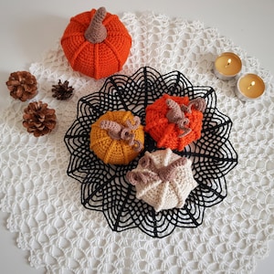 May include: Four crocheted pumpkins in orange, yellow, and white on a white crocheted doily with black lace trim. Two small candles are on the doily.