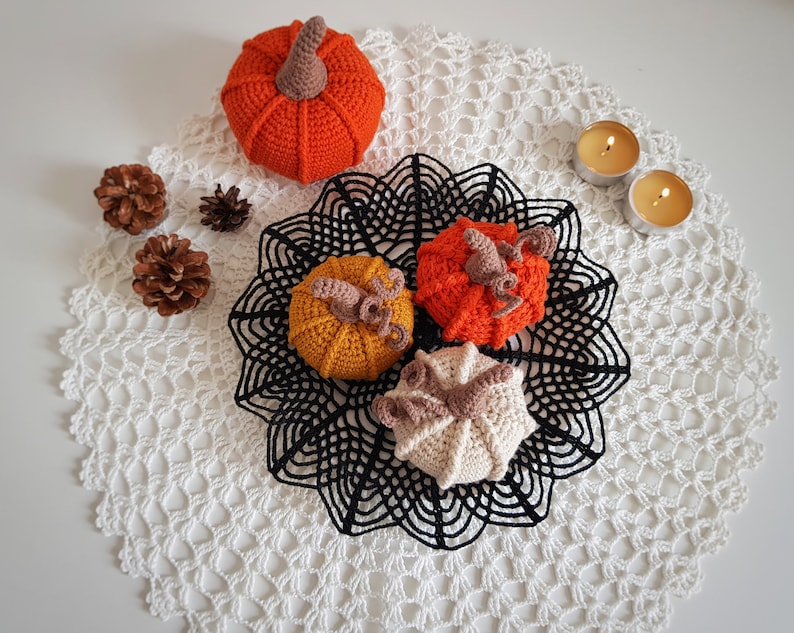 May include: Four crocheted pumpkins in orange, yellow, and white on a white crocheted doily with black lace trim. Two small candles are on the doily.