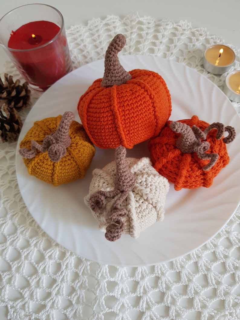 May include: Four crocheted pumpkins in different colors, orange, yellow, white and brown, on a white plate with a white crocheted doily underneath.