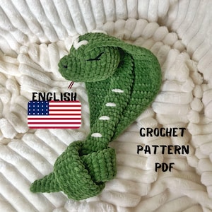 Puede incluir: Juguete de amigurumi de serpiente verde tejido a crochet con detalles blancos. El juguete está sobre una superficie blanca y esponjosa. El texto "ENGLISH" y una bandera de Estados Unidos son visibles en la imagen. El texto "CROCHET PATTERN PDF" también es visible.