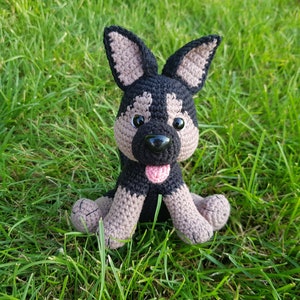 K&ouml;nnte beinhalten: Ein geh&auml;keltes Stofftier-Hund mit schwarzem und braunem Fell, braunen Augen, einer schwarzen Nase und einer rosa Zunge. Der Hund sitzt auf einem Bett aus gr&uuml;nem Gras.