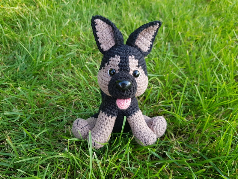 K&ouml;nnte beinhalten: Ein geh&auml;keltes Stofftier-Hund mit schwarzem und braunem Fell, braunen Augen, einer schwarzen Nase und einer rosa Zunge. Der Hund sitzt auf einem Bett aus gr&uuml;nem Gras.