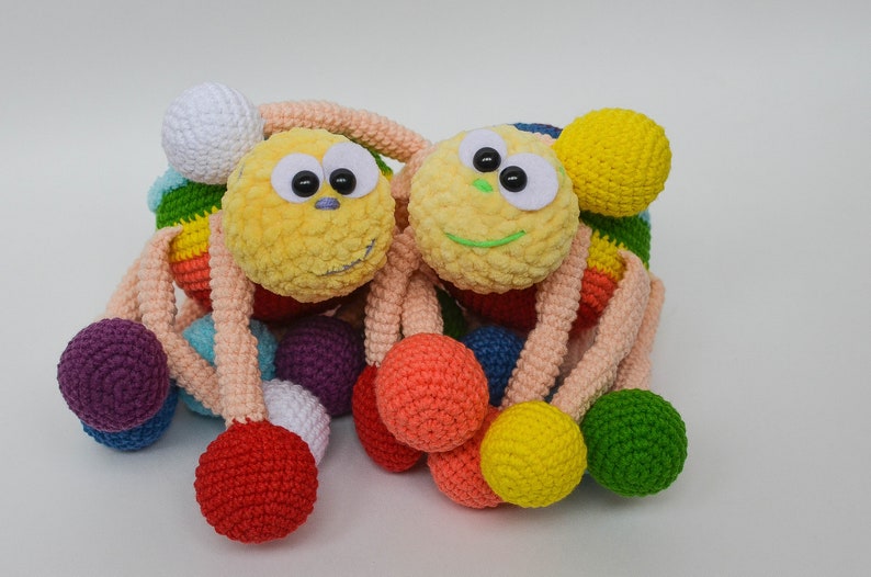 Pu&ograve; includere: Due giocattoli di peluche all'uncinetto a forma di ragno con palline colorate attaccate alle loro gambe. I ragni hanno facce gialle e bianche con occhi neri e sorrisi verdi.